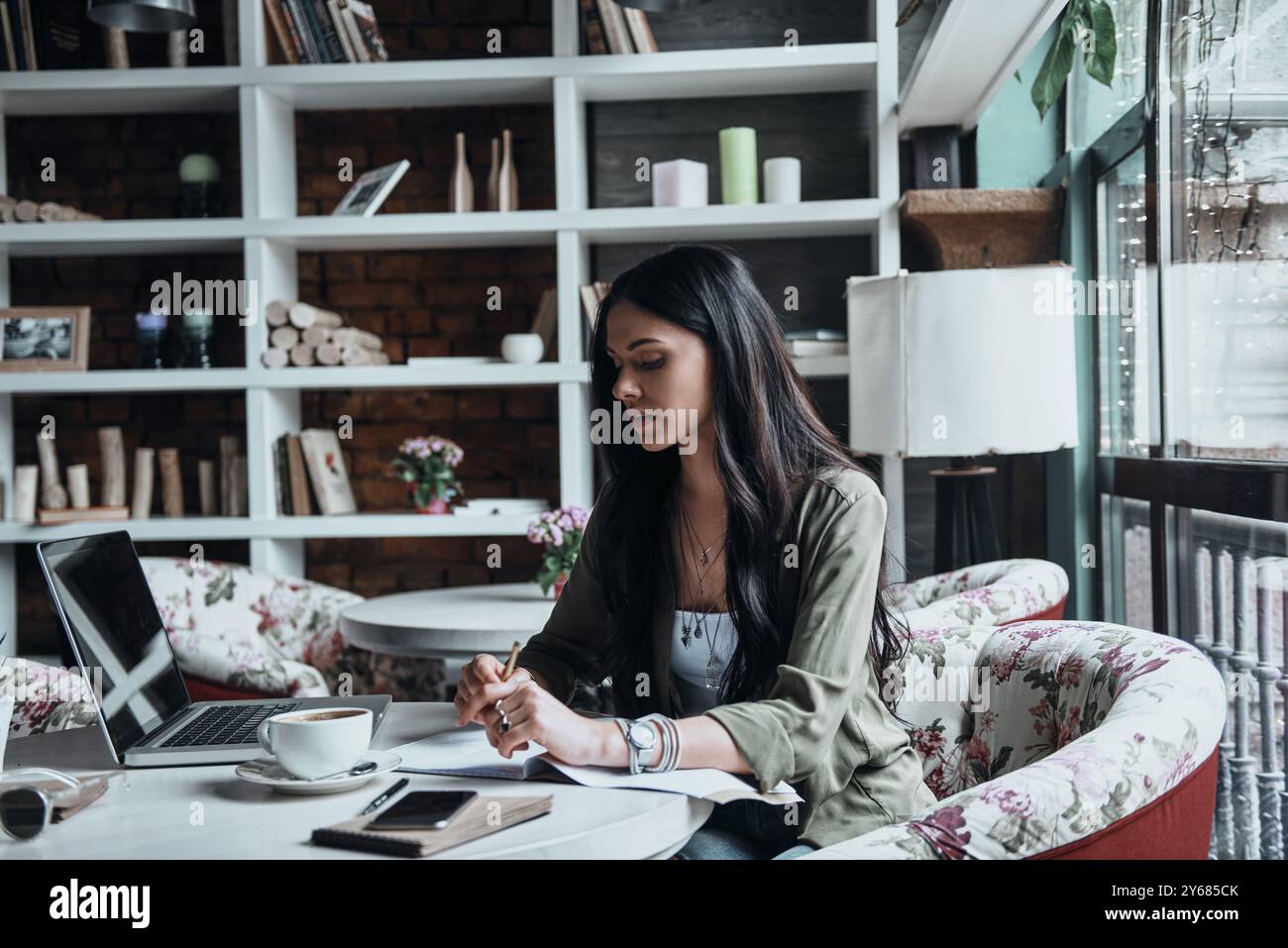 Sie schreibt ihre Gedanken auf. Eine nachdenkliche junge Frau schreibt etwas in ihr Notizbuch, während sie an ihrem Arbeitsplatz sitzt Stockfoto