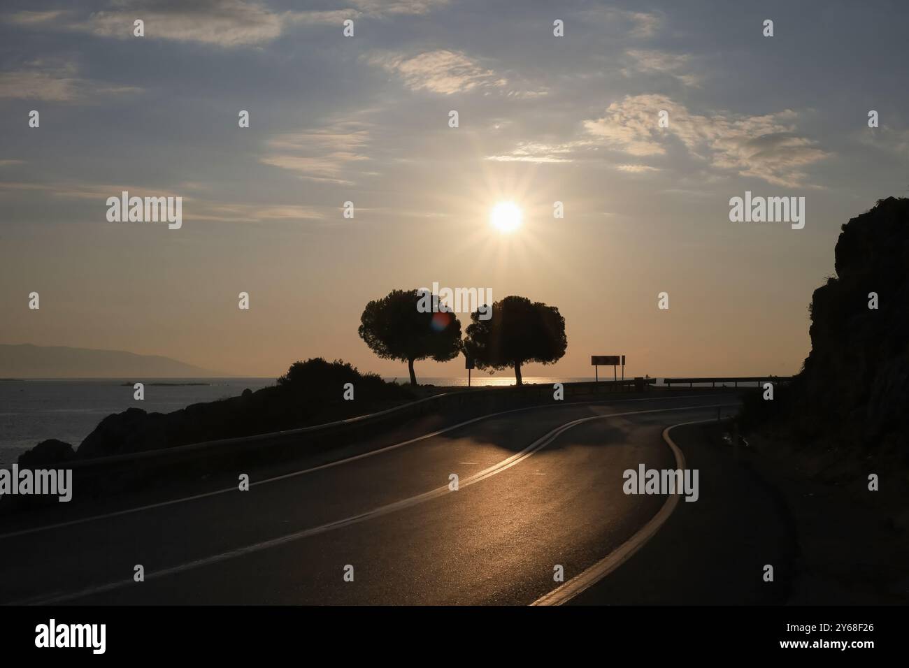 Ein malerischer Blick auf eine gekrümmte Straße, die zum Horizont führt, mit zwei Silhouettenbäumen unter der hellen untergehenden Sonne, die lange Schatten über den Rapp werfen Stockfoto
