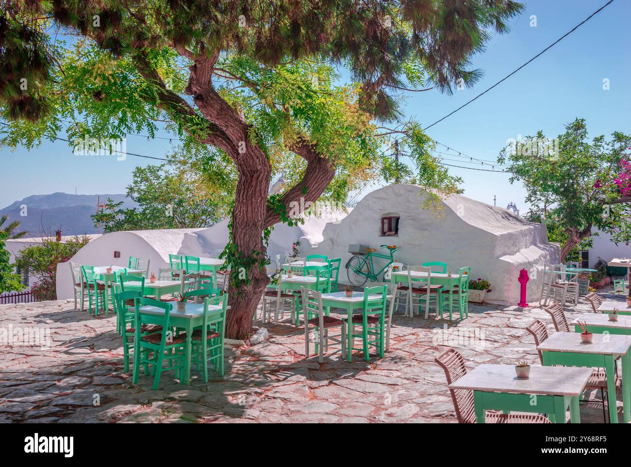 Café im Freien auf dem Platz von Hora, der Hauptstadt der Insel Amorgos, in Kykladen, Griechenland. Weiß getünchte Häuser und Tische im Schatten des Baumes. Stockfoto