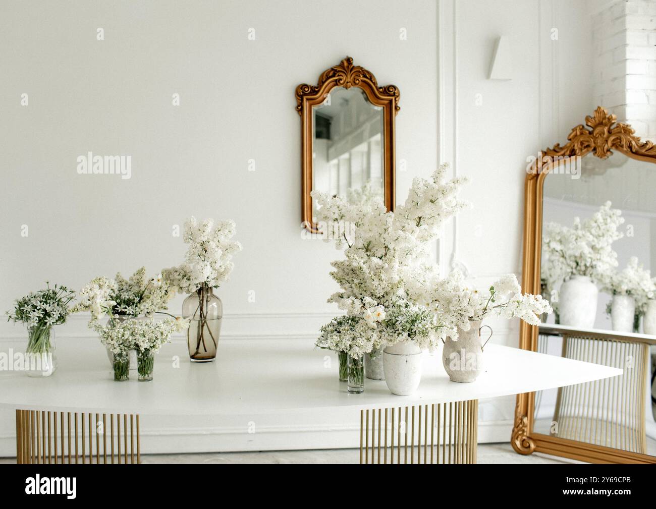 Helle weiße Blumen in verschiedenen Vasen schmücken einen stilvollen Esstisch. Vintage-Spiegel spiegeln die Eleganz des minimalistischen Dekors wider und schaffen eine ruhige Atmosphäre Stockfoto