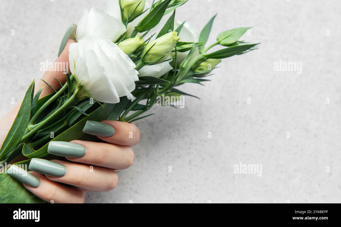 Eine Hand mit stilvollen langen Nägeln hält einen Blumenstrauß aus frischen weißen Blumen. Die sanften Grün- und Weißtöne bilden einen schönen Kontrast zu einem leicht strukturierten Rücken Stockfoto