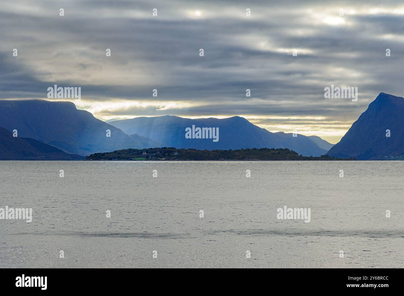 Ruhiges Wasser fängt das sanfte Leuchten der Dämmerung ein und offenbart eine einsame Insel, die von majestätischen Bergen und einem bewölkten Himmel umgeben ist. Stockfoto