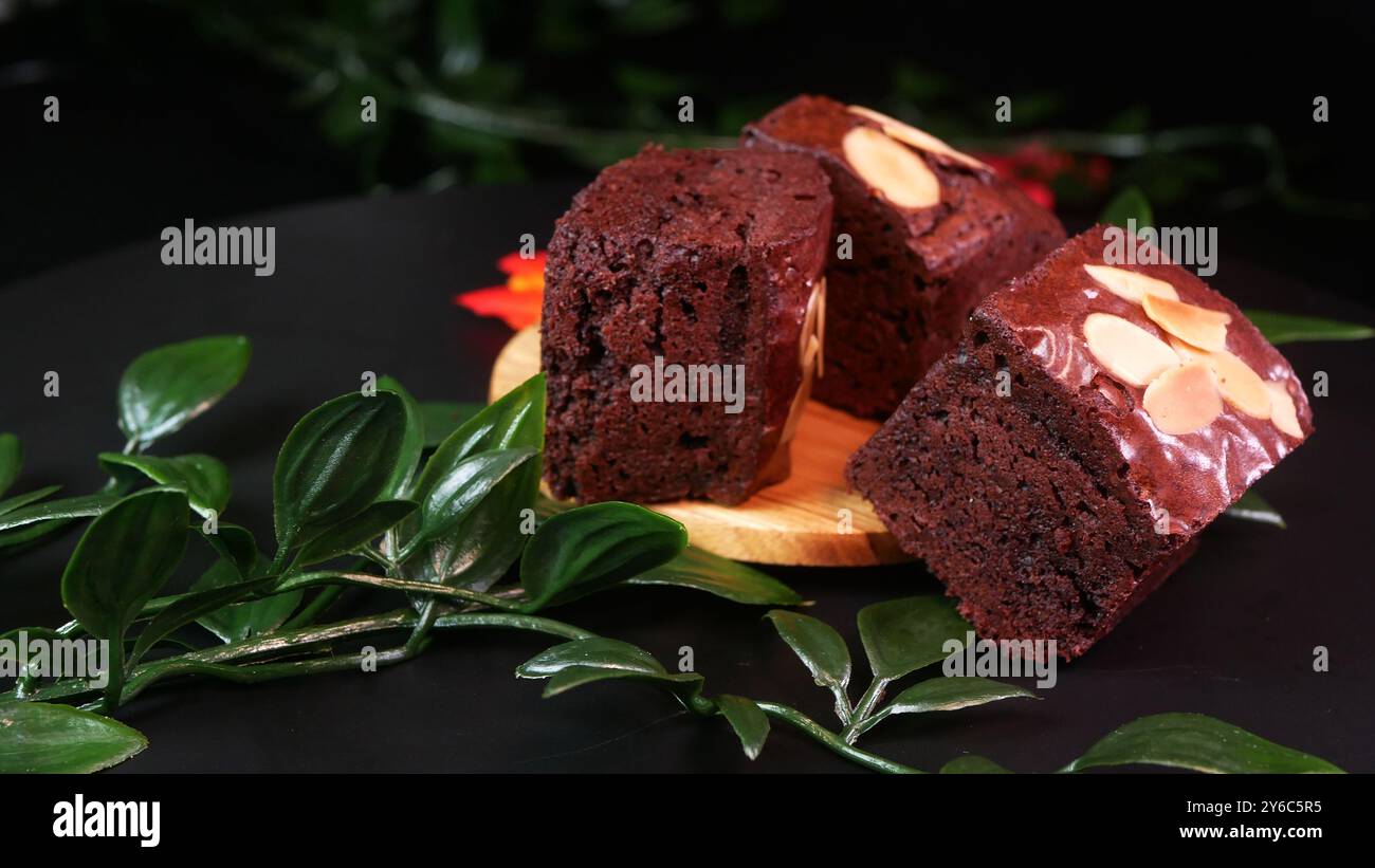 Weiche Schokoladen-Brownie-Stücke mit knusprigen Mandelbelag, serviert auf einem Holztablett mit grünen und roten Blattdekorationen, um ein natürliches Ambiente zu schaffen. Stockfoto