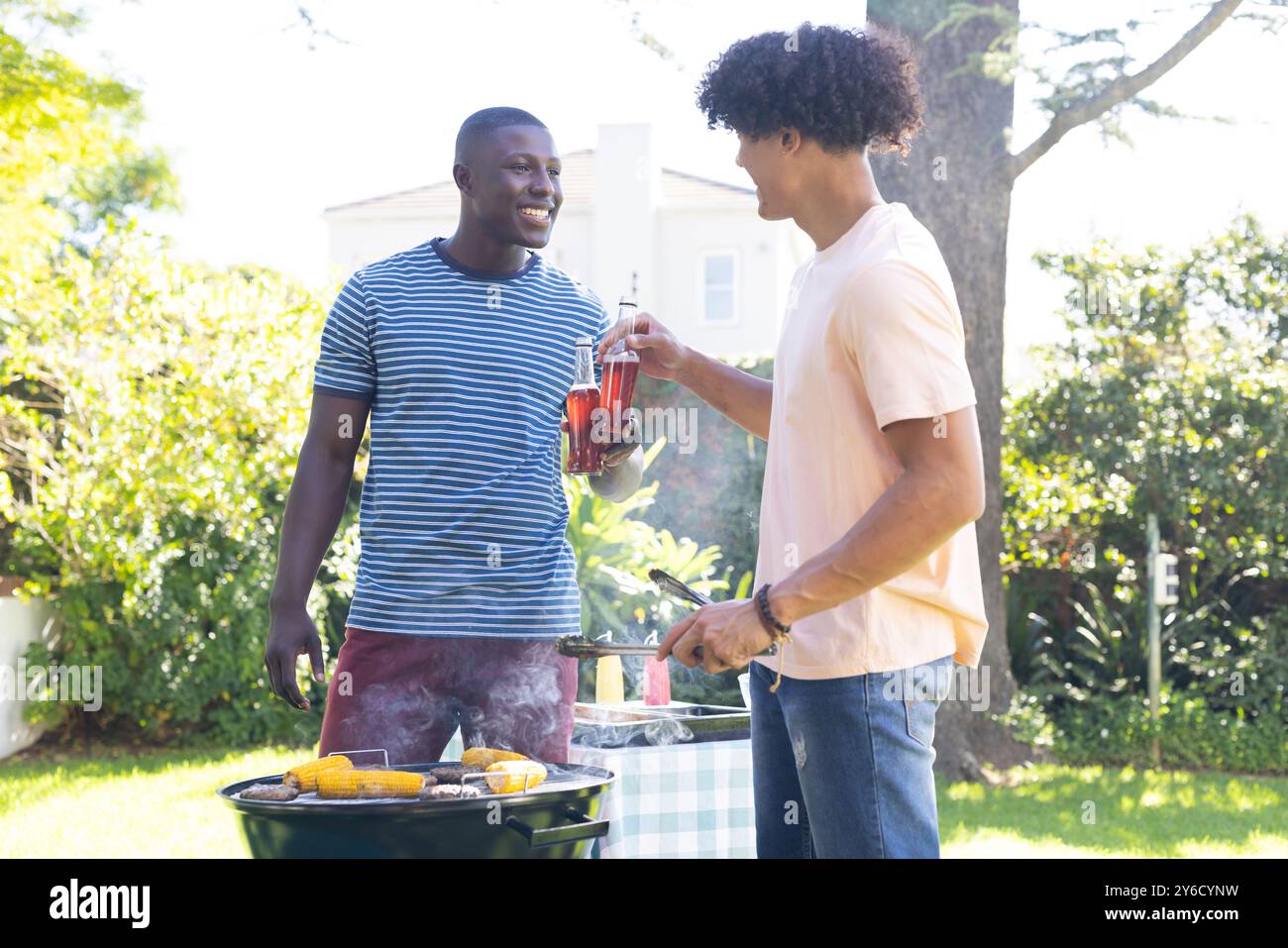 Grillen Sie Mais auf dem Grill, zwei verschiedene männliche Freunde genießen Drinks und plaudern Sie im Freien Stockfoto