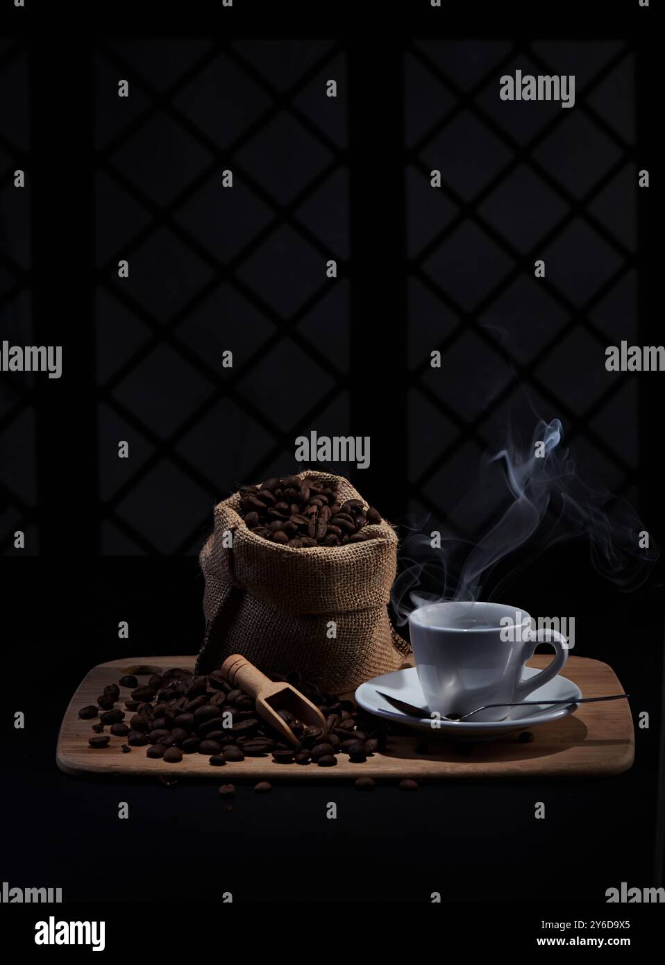 Kaffeebohnen, kleiner Kaffeelöffel, Kaffeetasse mit dünnem Rauch von heißem Kaffee, fotografiert auf einem Tablett mit Fensterschatten Hintergrund Stockfoto