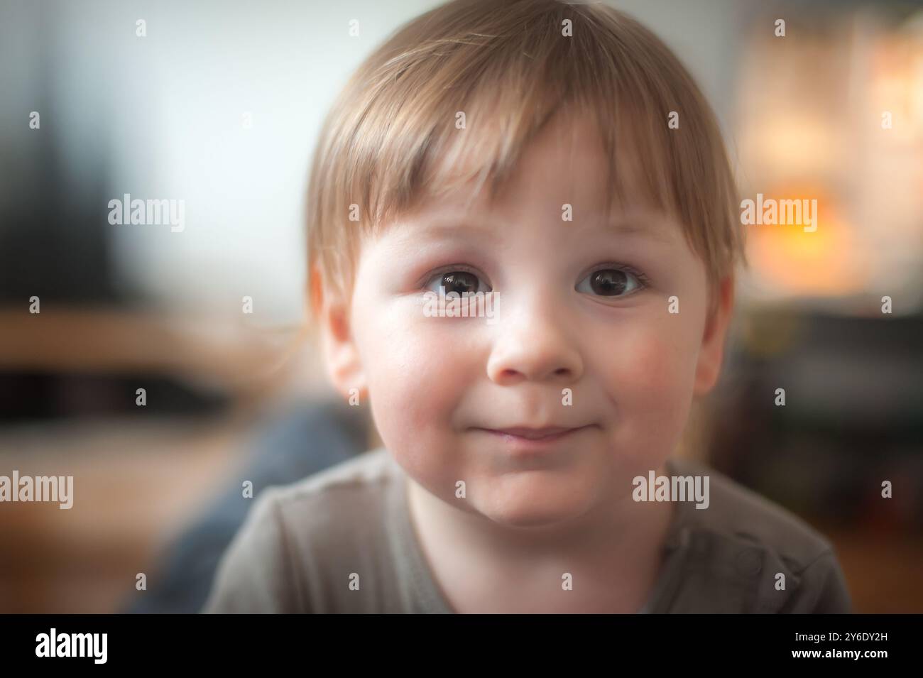 Ein fröhliches kleines Kind mit ausdrucksstarken Augen strahlt vor Glück, während es gemütlich drinnen sitzt. Das Ambiente strahlt eine warme und einladende Atmosphäre aus, Stockfoto
