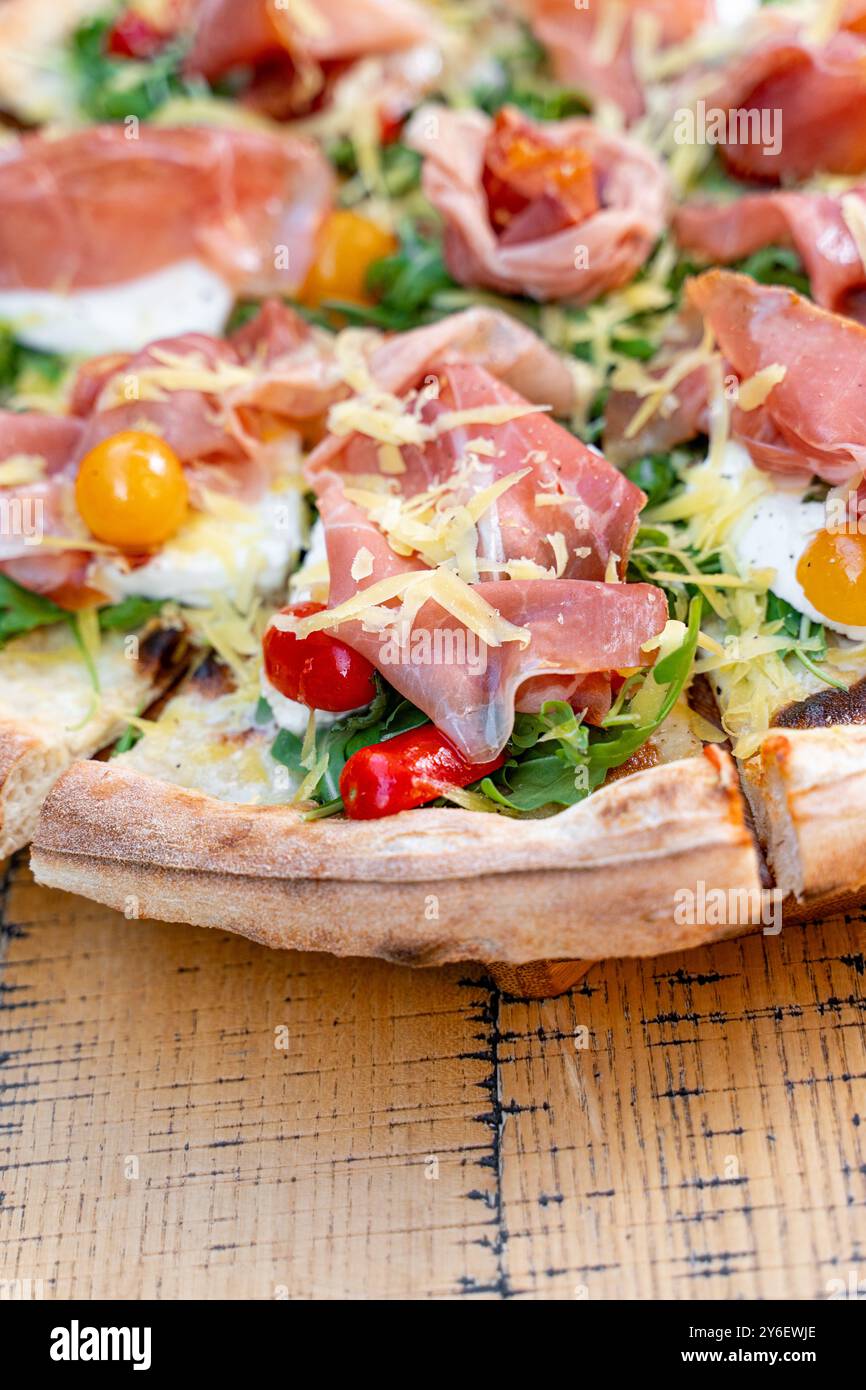 Nahaufnahme der handwerklichen Pizza mit Schinken, frischem Rucola, bunten gelben und roten Kirschtomaten und geriebenem Käse. Serviert auf rustikalem Holz Stockfoto