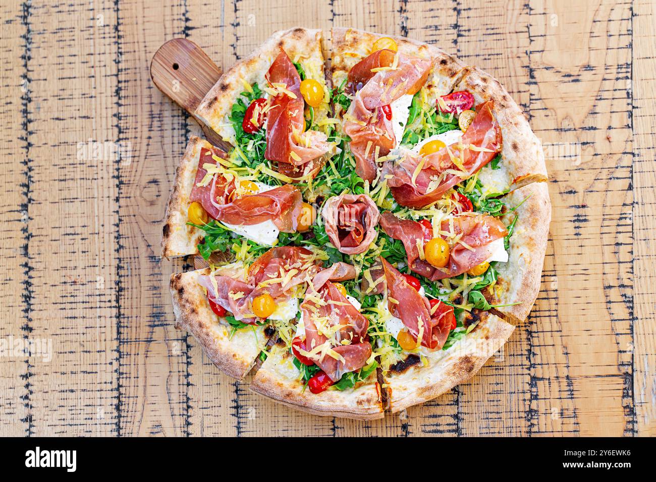 Handwerkliche ganze Pizza mit Schinken, frischem Rucola, gelben und roten Kirschtomaten und geriebenem Käse. Serviert auf einem rustikalen Holzbrett, Showcas Stockfoto
