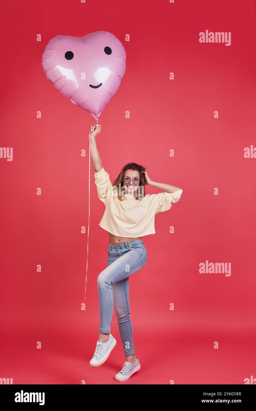 Lass die Party beginnen! Die ganze Länge einer attraktiven jungen lächelnden Frau, die einen herzförmigen Ballon hält und die Hand im Haar hält, während sie gegen pi steht Stockfoto