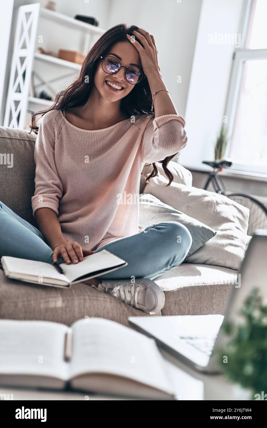 Untersuchungsvorbereitung. Schöne junge Frau in Brillen, die studiert und lächelt, während sie zu Hause auf dem Sofa sitzt Stockfoto