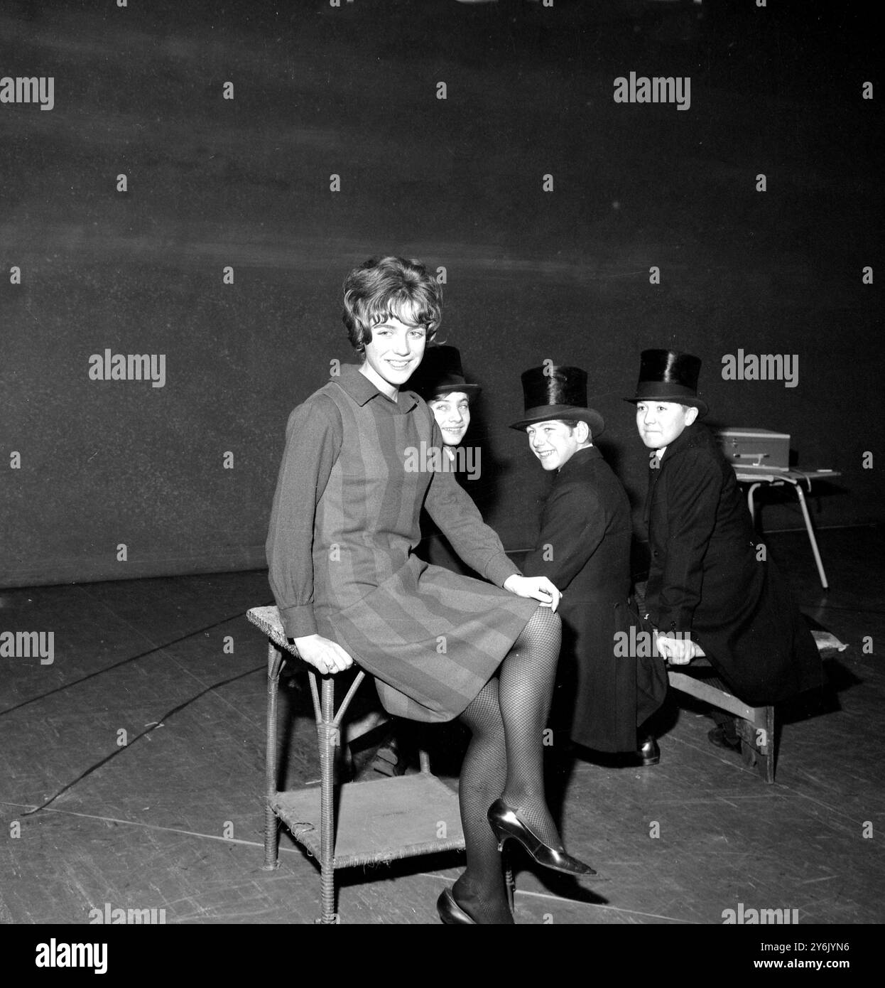 Die Schauspielerin Kika Markham kommt bei den Proben in London für das Stück " The Fourth of June " unter die Augen dieser drei jungen Etonier , die Bühnenfassung des Bestsellerromans über Eton des alten etonischen David Benedictus . Das Stück wurde vor london im Theatre Royal Brighton gespielt. Die drei jungen Jungs sind ( von links nach rechts ) Kit Williams , Paul Holdaways und John Chipperfield . 9. Januar 1964 Stockfoto