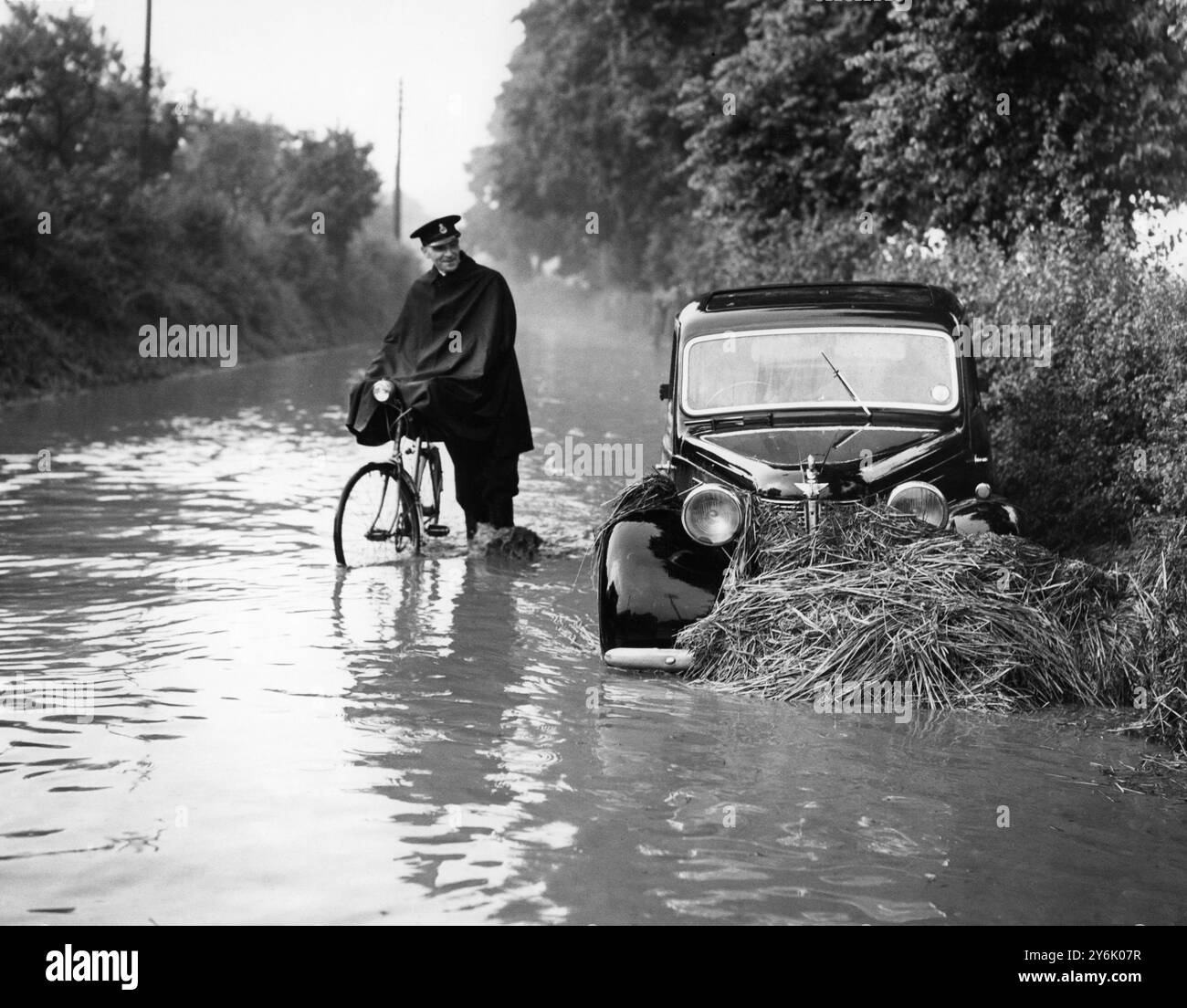 Mr. John Martindale, der dieses Auto in Sutton in Hone fuhr, in der Nähe von Dartford, Kent, wurde heute Abend von Flood Water gestürzt. Als er aus dem Auto stieg, brach ihm das Wasser mit Ziegelschutt beide Beine. Ein Mann auf einem Fahrrad in Uniform sieht sich die Schäden an, die durch die Überschwemmung verursacht wurden. 1956 Stockfoto