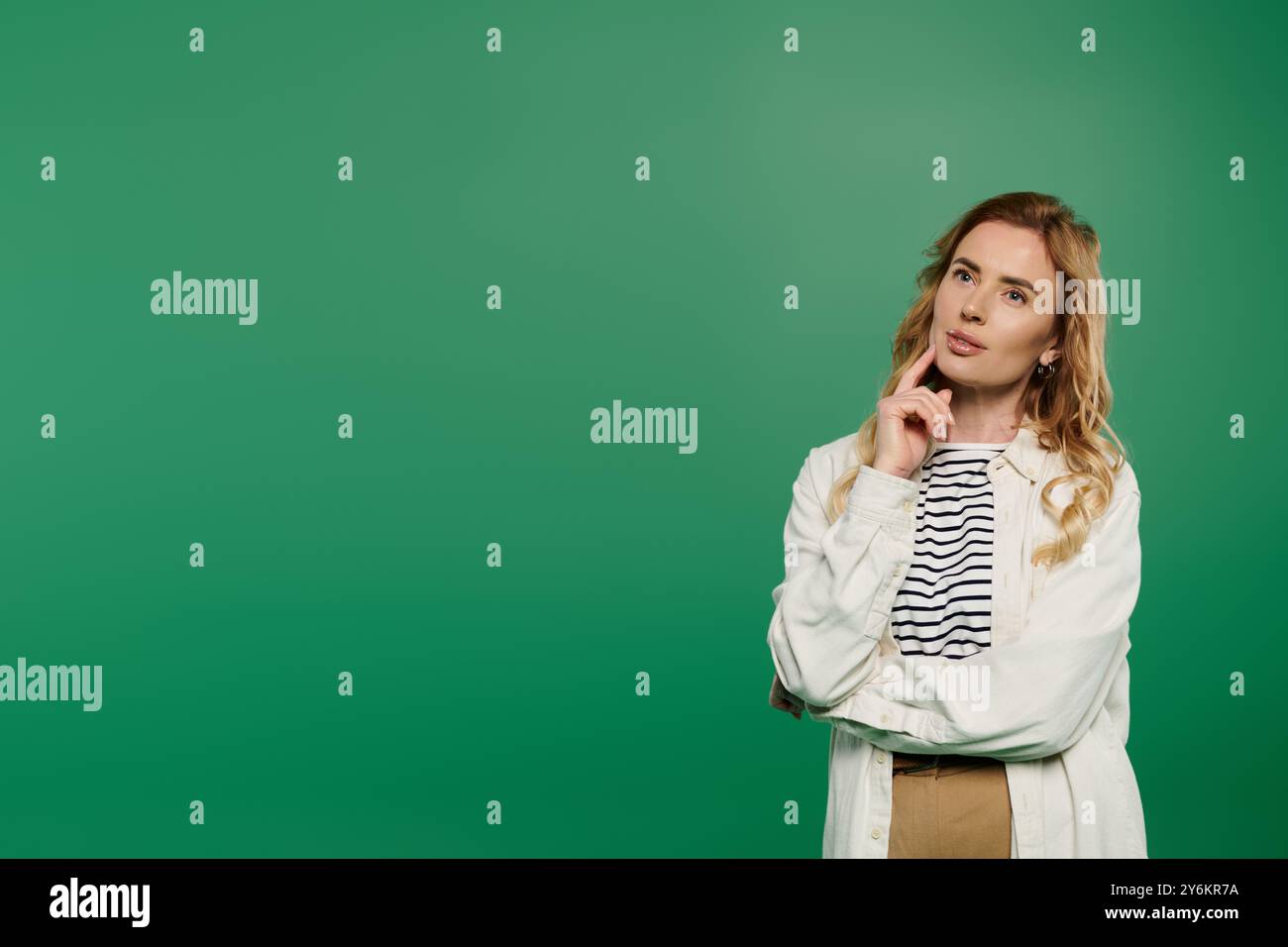 Eine nachdenkliche Frau posiert mit einem Finger auf dem Kinn und zeigt legeren Style und lebhafte Locken vor grünem Hintergrund. Stockfoto