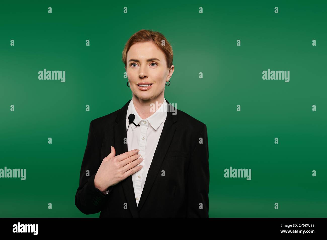 Ein professioneller Fernsehmoderator in eleganter schwarzer Kleidung spricht ein Publikum vor einem leuchtend grünen Hintergrund an. Stockfoto