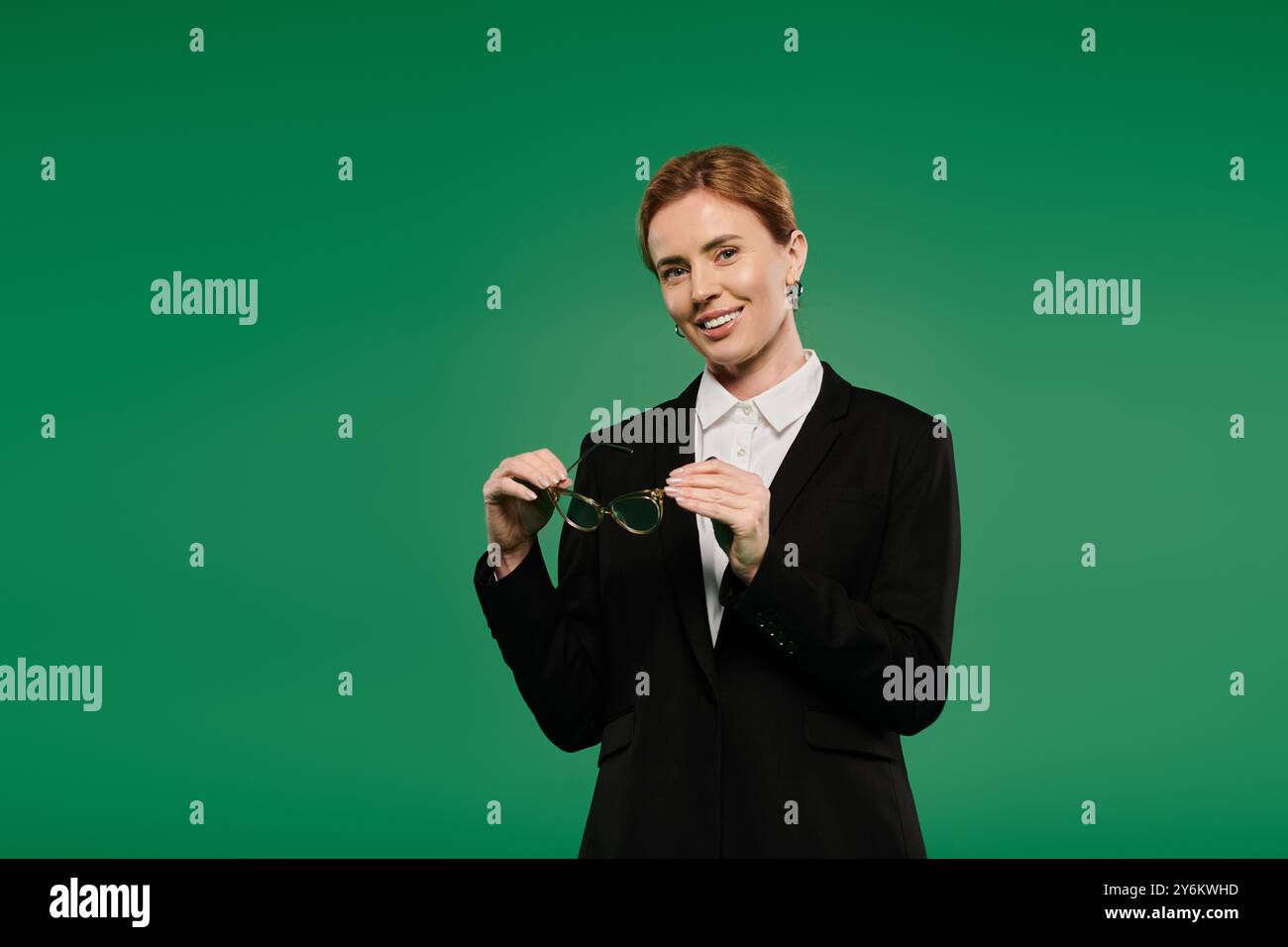 In formellem Schwarz gekleidet, zeigt ein Fernsehmoderator ein warmes Lächeln, während er die Brille vor einem leuchtend grünen Hintergrund hält. Stockfoto