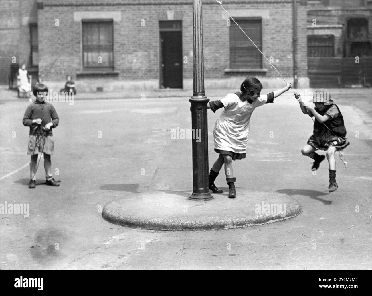 1930 s (?) Mädchen spielen auf der Straße ein Spiel, schwingen an einem Seil, das an einem Lampenpfosten gebunden ist. Zwei spielen am Seil, während ein anderes Mädchen im Hintergrund zusieht und ein Seil hält. Zwei ist Gesellschaft, drei ist eine Menge Stockfoto