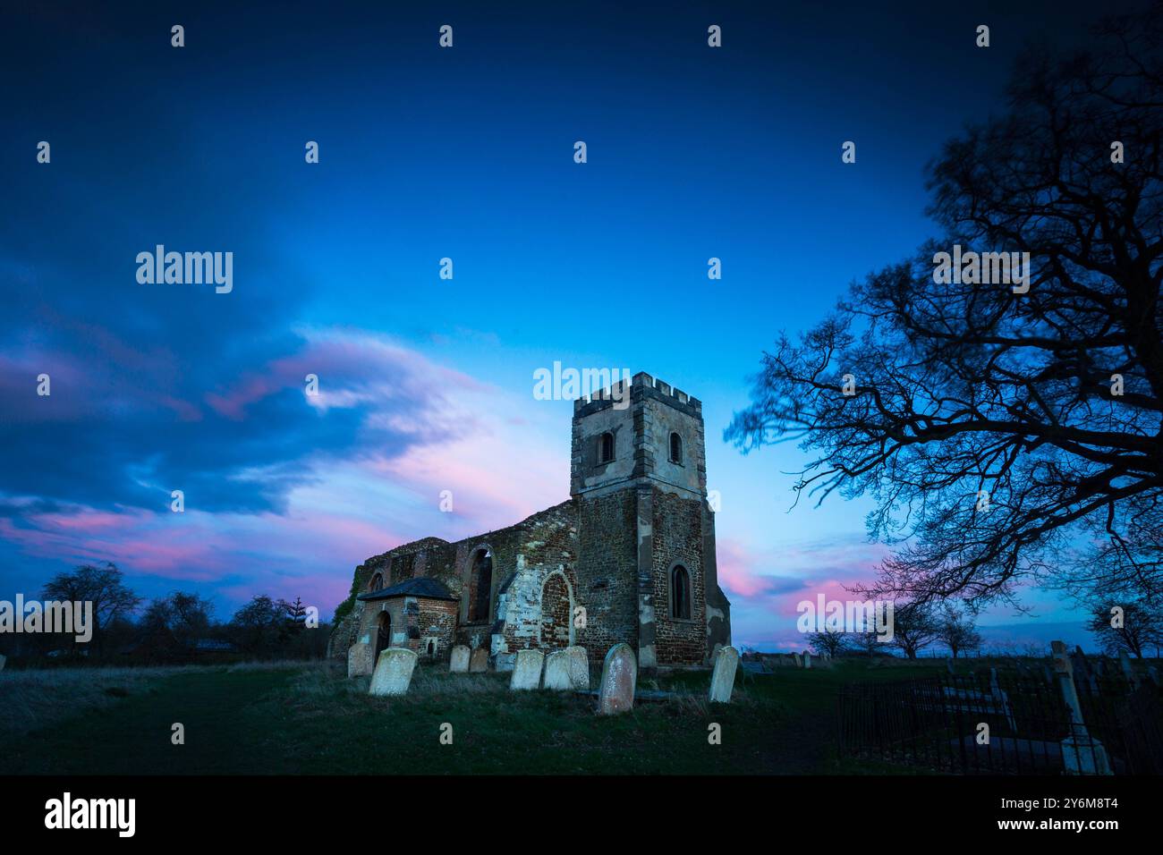 Eine alte Kirchenruine mit Grabsteinen unter einem lebendigen Himmel bei Sonnenuntergang, mit Silhouettenbäumen im Hintergrund. Stockfoto