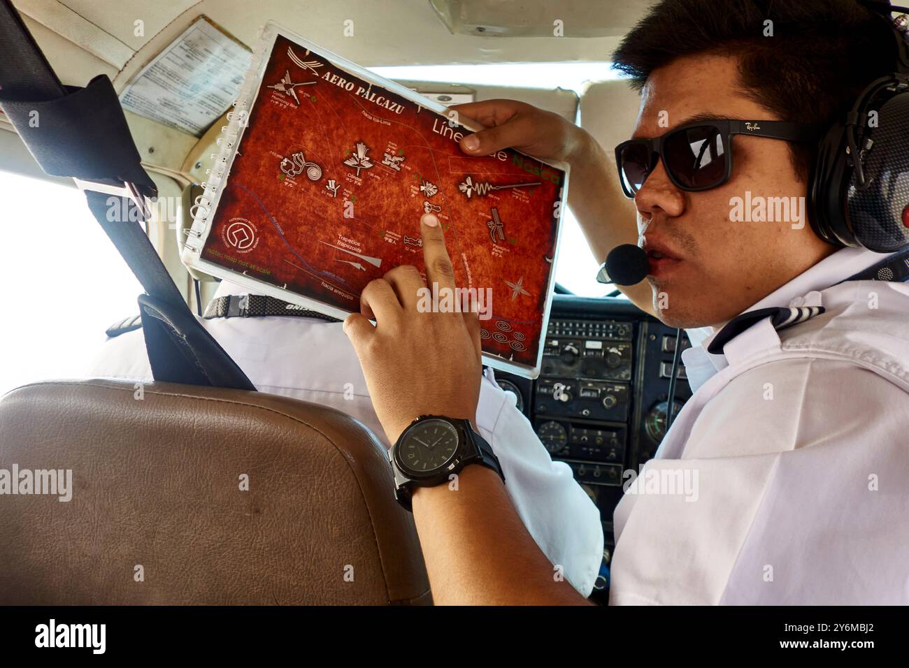 Ein Pilot mit Sonnenbrille und Kopfbedeckung zeigt auf einer Karte der Nazca-Linien, die die Passagiere während einer Luftfahrt begleitet. Die Cockpit-Einstellung verbessert die AVI Stockfoto