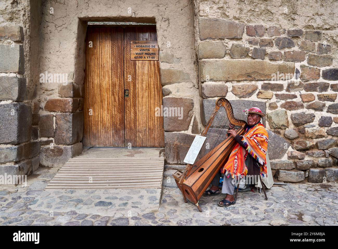 Ein traditioneller Musiker in farbenfroher Kleidung spielt Harfe in den historischen Straßen von Ollantaytambo, Peru. Die Szene fängt das kulturelle Erbe und l Stockfoto