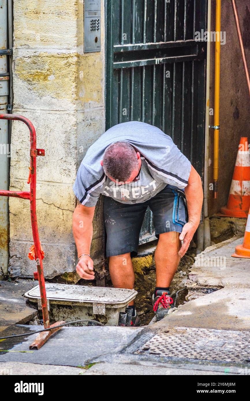Arbeiter in Shorts, die sich nach unten beugen und die Reparaturarbeiten im Loch im Boden betrachten - Tours, Indre-et-Loire (37), Frankreich. Stockfoto