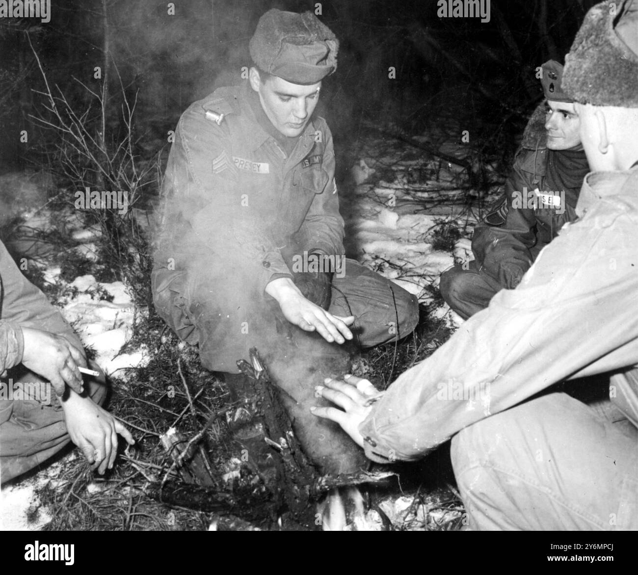 Grafenwoeur, Deutschland: Rock 'n Roll Sänger Elvis Presley, heute kommissarischer Sergeant in der US-Armee, wärmt sich hier am 6. Februar in seiner letzten Nacht auf Manövern die Hände vor einem rauchigen Feuer, nachdem er an den gemeinsamen amerikanisch-deutschen Armee-Wintermanövern teilgenommen hatte, die am 7. Februar endete. Presley wird sich bald darauf vorbereiten, nach Ablauf seiner zwei Dienstjahre aus der Armee wieder in die Vereinigten Staaten zu entlassen. 8. Februar 1960 Stockfoto