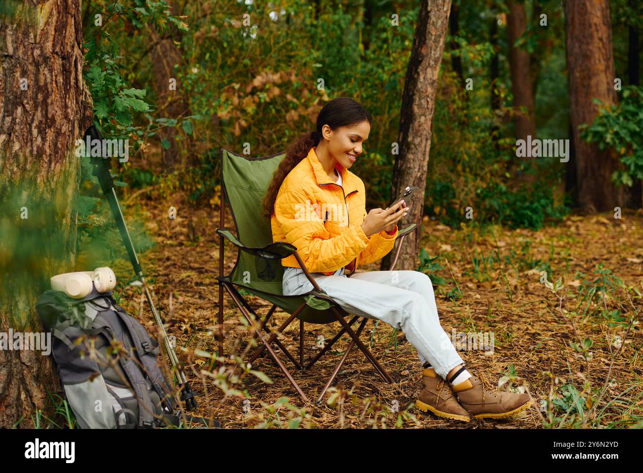 Eine junge Afroamerikanerin entspannt sich im Herbstwald und erkundet ihr Telefon inmitten lebendigen Laubs. Stockfoto