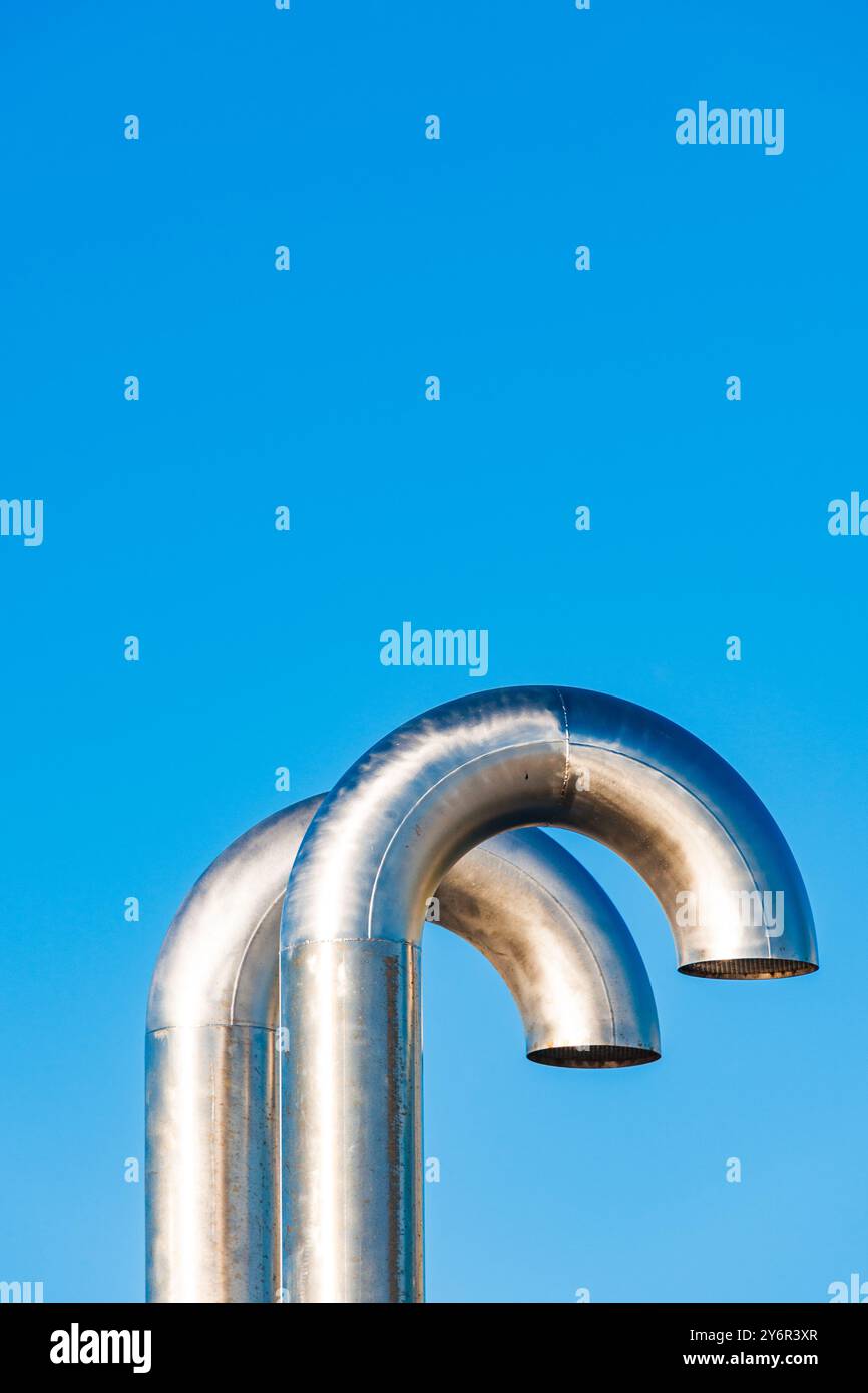 Zwei glänzende, geschwungene Metallrohre heben sich von einem leuchtend blauen Himmel ab und zeigen ein industrielles Design. Die Szene spiegelt eine moderne technische Ästhetik wider Stockfoto