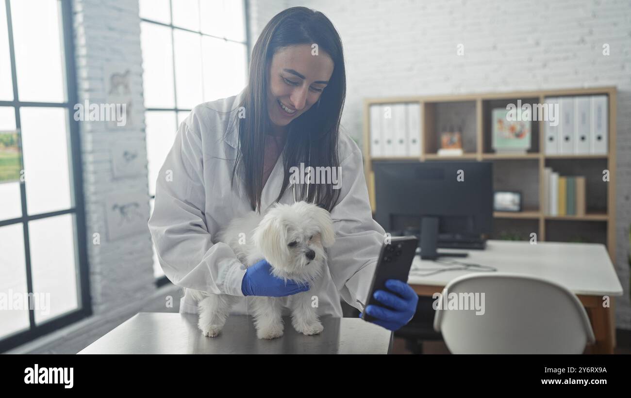 Eine lächelnde Tierärztin in einem weißen Mantel hält einen maltesischen Hund auf einem Untersuchungstisch, während sie ihr Handy in einer Klinik ansieht. Stockfoto
