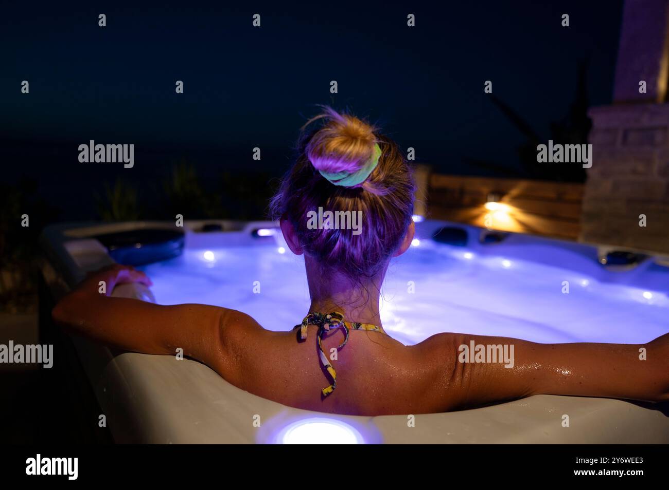 Blick von hinten auf eine Frau, die an einem Sommerabend draußen einen Whirlpool genießt. Mit blauen Lichtern im Whirlpool. Gemütliche und luxuriöse Auszeit. Stockfoto