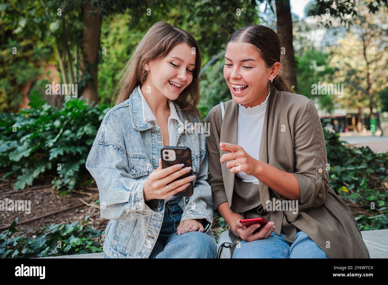 Zwei junge Frauen sitzen draußen, lächeln und interagieren mit Inhalten auf einem Mobiltelefon, nutzen Smartphones, um spannende Momente zu teilen, indem sie die digitale Kommunikation und die Interaktion in sozialen Medien hervorheben. Hochwertige Fotos Stockfoto