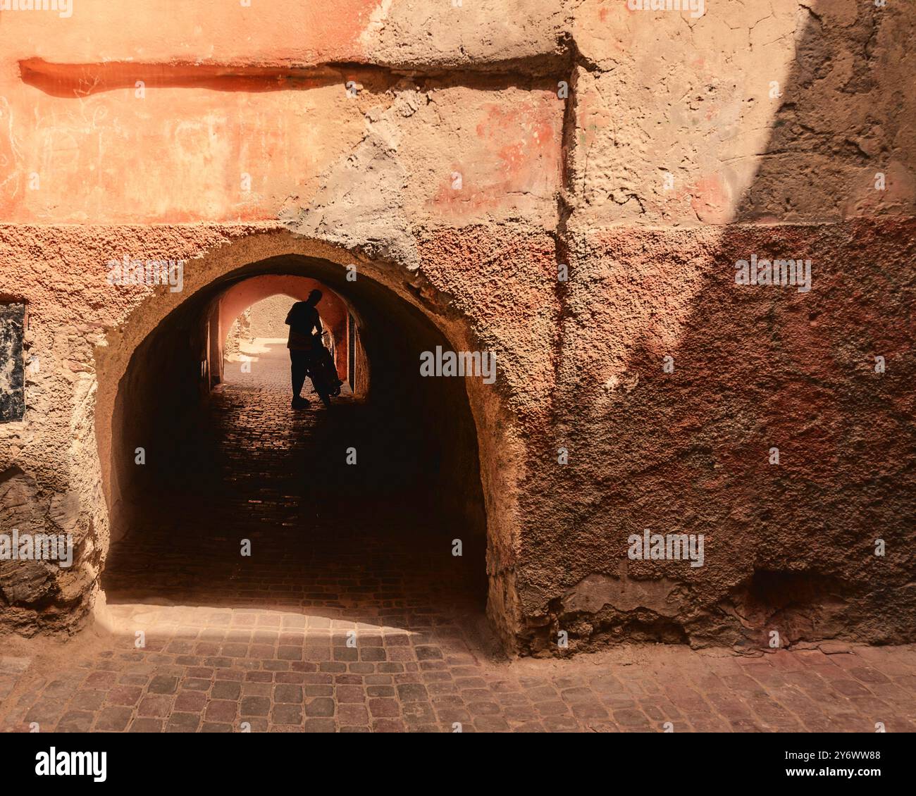 Eine Silhouette einer Person, die mit dem Fahrrad durch einen bogenförmigen Durchgang in einer strukturierten, verwitterten Straße im alten Marrakesch in der Medina, Marokko, geht. Stockfoto