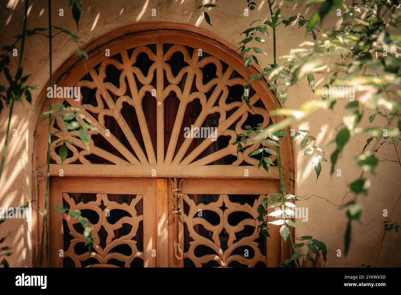 Kunstvolle marokkanische traditionelle Holztür mit kompliziertem Gitterdesign, strukturierter Wand und umliegendem Grün in einem ruhigen, luxuriösen Riad-Innenhof mit Stockfoto