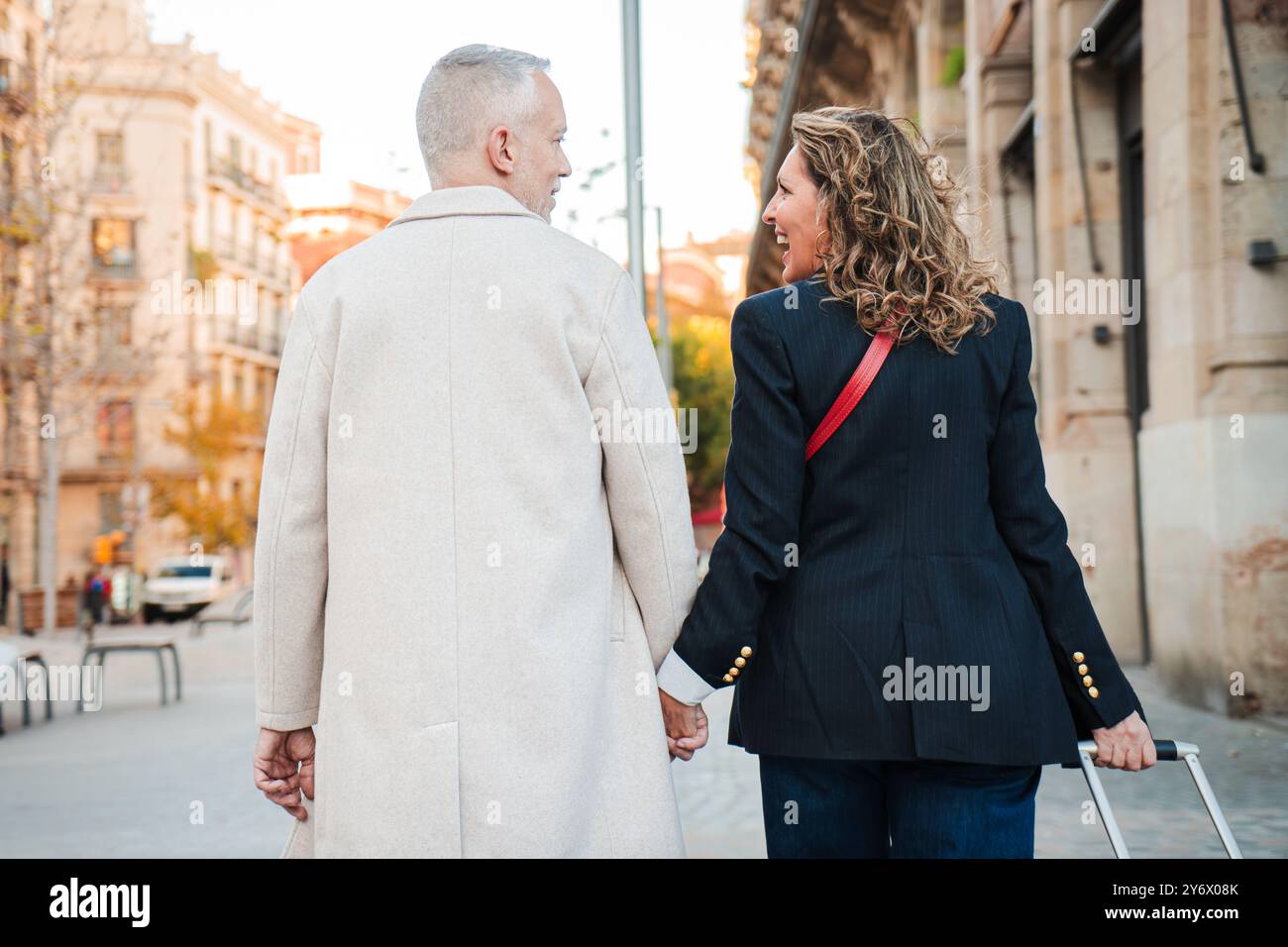 Reife Ehepaare gehen Hand in Hand durch eine Stadtstraße, genießen einen entspannten Spaziergang zusammen, während sie ihr Gepäck ziehen, Liebe und Gesellschaft zeigen und tagsüber in einer urbanen Umgebung reisen. Hochwertige Fotos Stockfoto