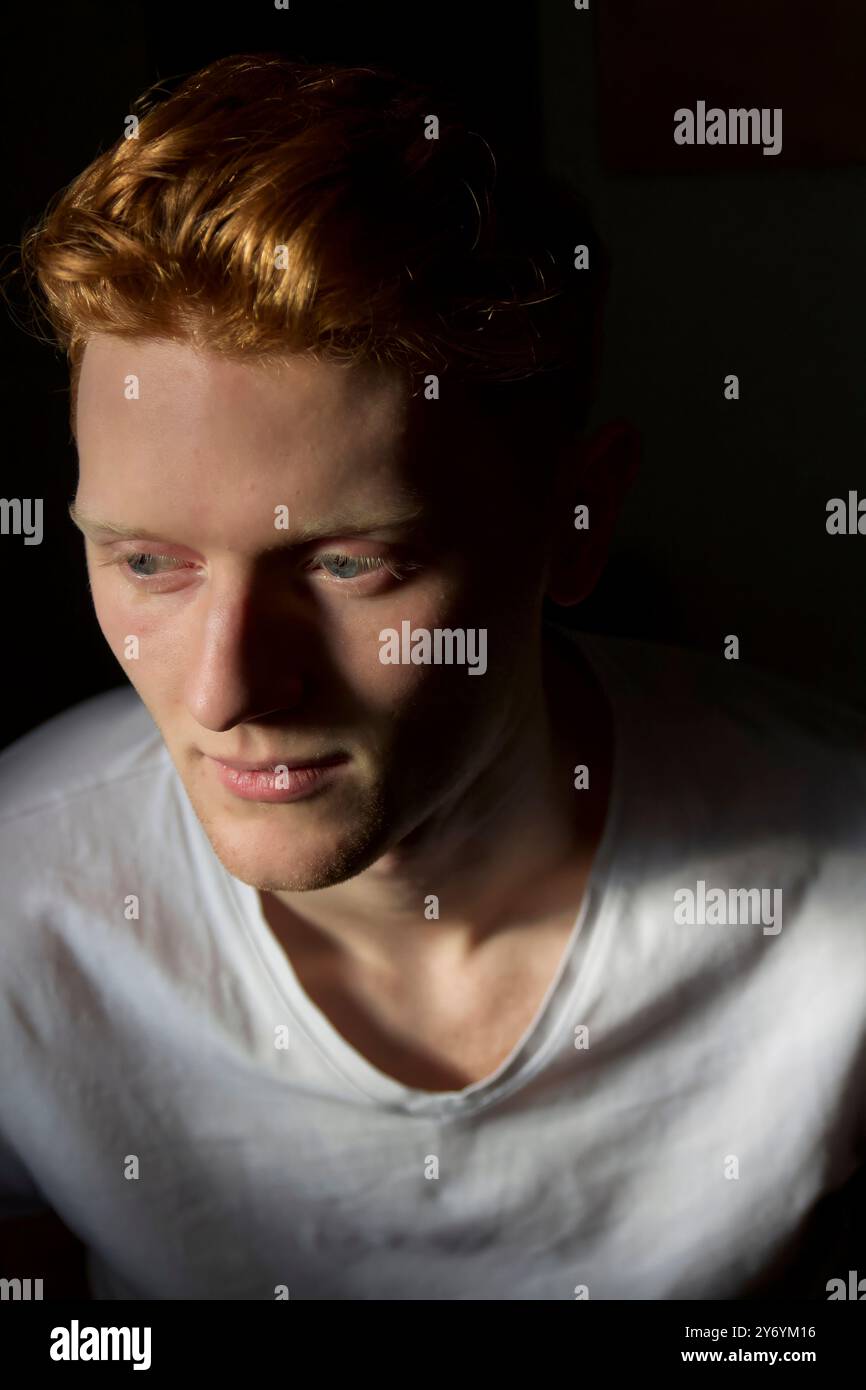 Junger Mann mit dunkelbraunem Haar, der in dunklem Licht sitzt, während er tief nachdenklich ist Stockfoto