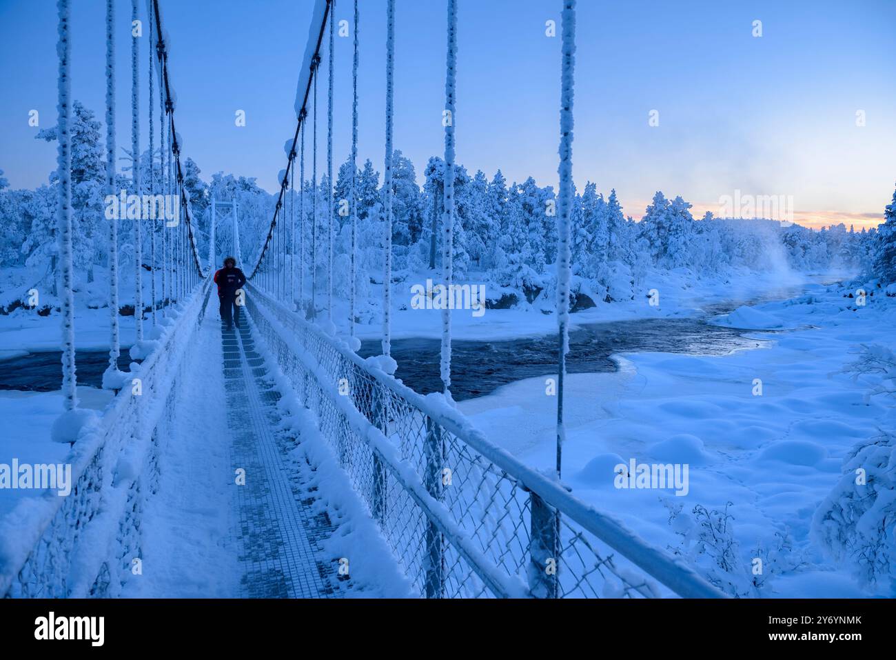 Hängebrücke über den verschneiten und gefrorenen Juutuanjoki-Fluss bei einem kalten Winteruntergang und Dämmerung in Inari (Lappland, Finnland) Stockfoto