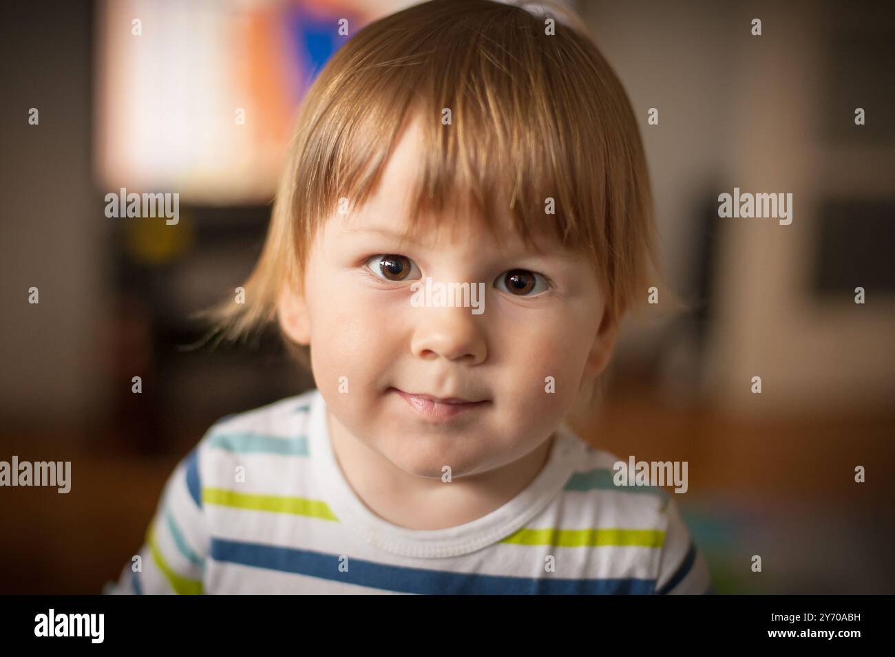 Ein kleines Kind mit hellbraunen Haaren und einem fröhlichen Ausdruck steht drinnen, trägt ein gestreiftes Hemd und lenkt die Aufmerksamkeit auf bunte Elemente im Th Stockfoto