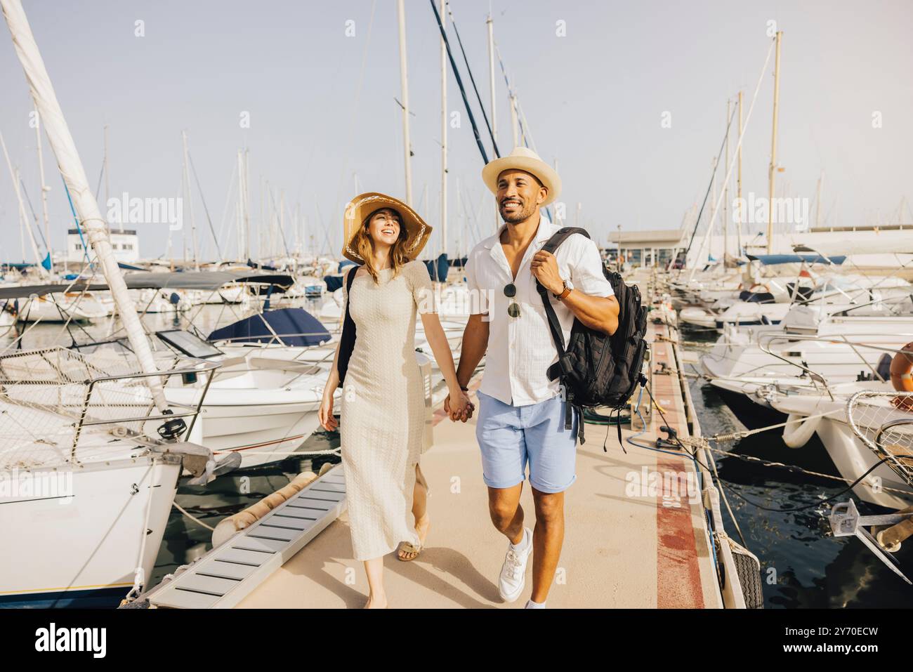 Ein lächelndes Paar spaziert Hand in Hand am Dock des Yachthafens entlang, umgeben von Booten an einem sonnigen Tag. Stockfoto