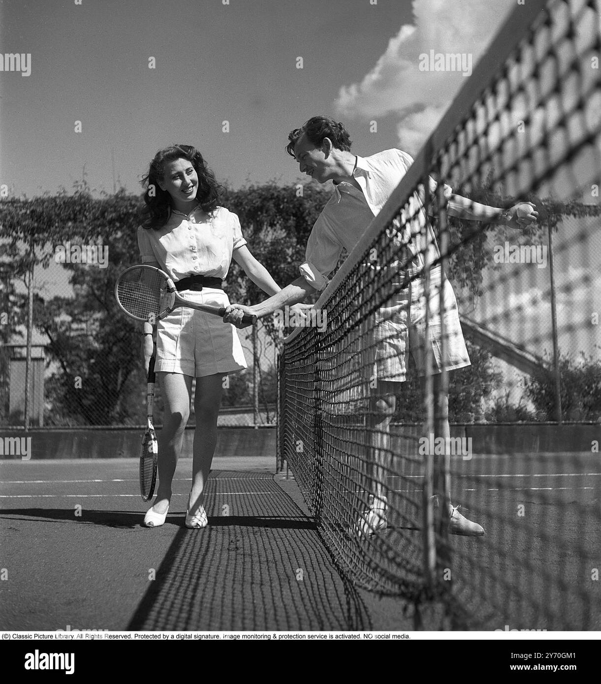 Ein Paar auf dem Tennisplatz 1945. Ein Paar auf dem Tennisplatz, das dieses Jahr Tennis-Mode-Kleidung trägt. Er hält seinen Schläger aus, als würde er zeigen, wie der Vorhandschlag gemacht wird. Kristoffersson Ref. O127-3 Stockfoto