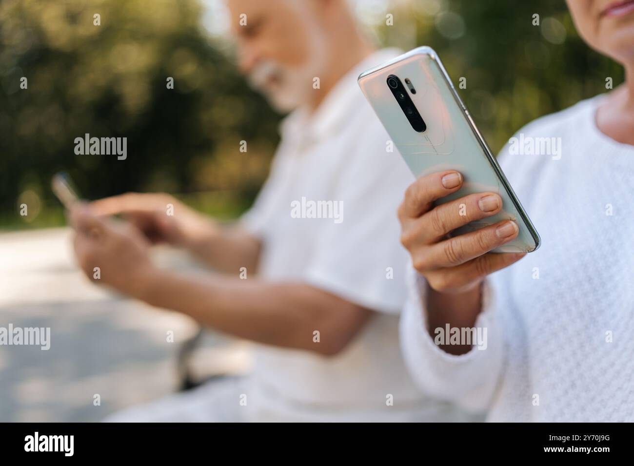 Close-up Porträt eines schönen älteren Paares mit Smartphones, die an sonnigen Sommertagen auf der Parkbank sitzen und in Verbindung bleiben Stockfoto