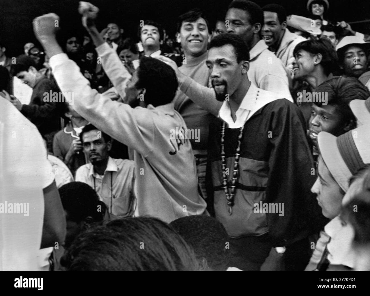 Der verbotene amerikanische Athlet John Carlos , dessen "Liebesperlen " an seinem Hals hängen , steht leise, während Mitglieder der jamaikanischen Olympischen Mannschaft geballte Fäuste - das Black-Power-Symbol - rufen und hochhalten , das Carlos und Teamkollege Tommie Smith bei ihrer Medaillenverleihung am 16 . Oktober gezeigt haben , um ihre Aussetzung von den 19. Olympischen Spielen herbeizuführen . Carlos war in der Menge im Olympiastadion , um anderen amerikanischen schwarzen Athleten bei verschiedenen Veranstaltungen zu zusehen . 19. Oktober 1968 Stockfoto
