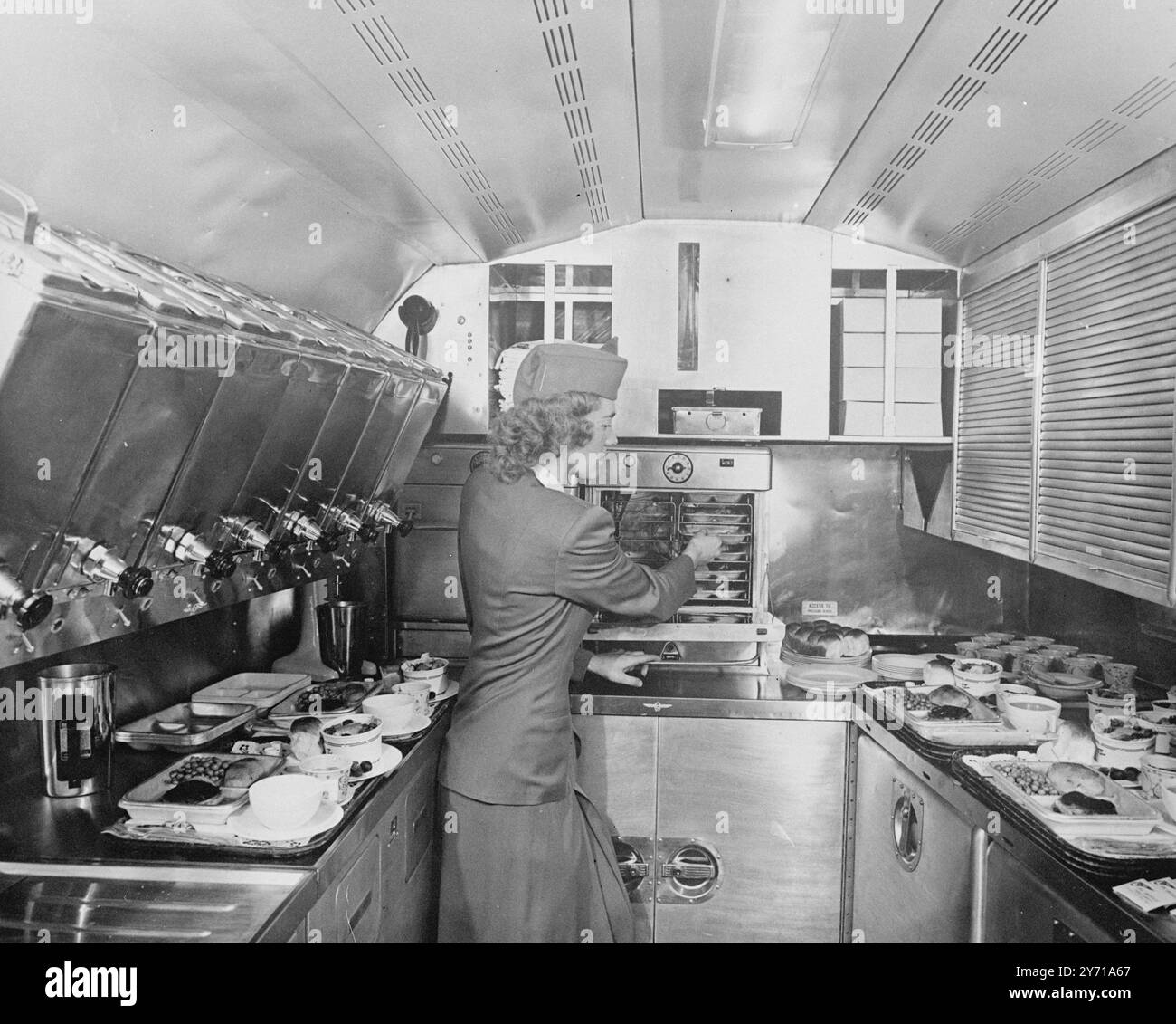 FLUGKÜCHE die Galerie an Bord von Pan American Airways New Clipper '' America '' kann 75 Passagiere und eine achtköpfige Besatzung dreimal bedienen , ohne aus einem Raum nachzuladen , der nicht viel größer ist als eine durchschnittliche Haushaltsküche . Alle Passagiere erhalten ihre Mahlzeiten innerhalb einer Stunde nach der ersten Portion . Sie werden Mahlzeiten essen, die im Ofen richtig zubereitet werden, nicht warme Lunchpakete. In der Galerie befinden sich zwei Öfen , einer oben offen gezeigt , mit einer Kapazität für jeweils 24 Mahlzeiten , vier Kühlschränke , sieben 6-Gallonen-Flüssigkeitsbehälter und nicht weniger als 15 Schubladen in verschiedenen Größen . Kaffee, fre Stockfoto