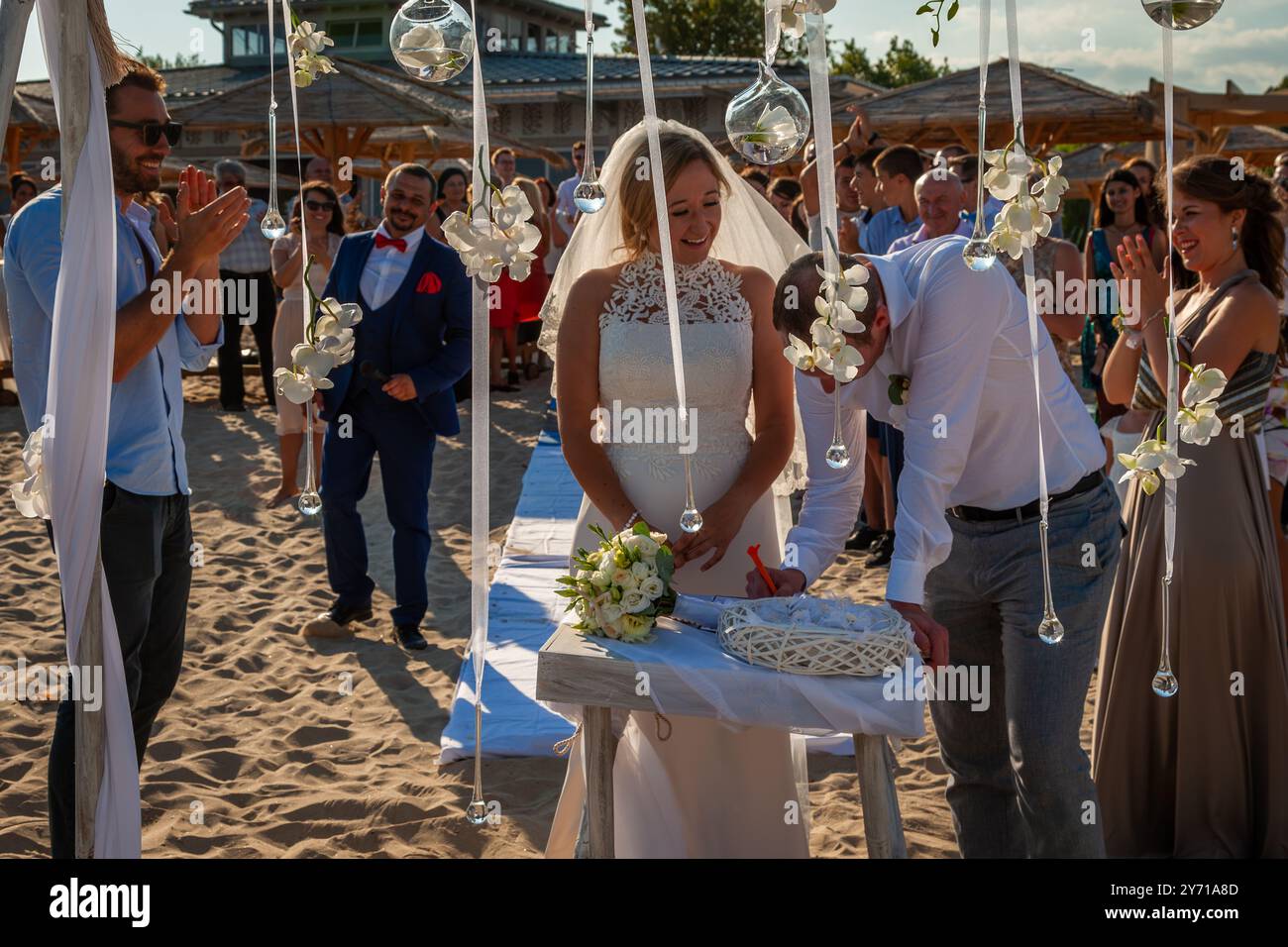 An einem kleinen Tisch, der mit Blumen und hängenden Glasbaubles geschmückt ist, unterschreibt der Bräutigam seinen Namen auf dem Heiratsurkunde, während applaudierende Gäste sich versammeln. Aufgenommen bei einer bulgarischen Strandhochzeit in Albena, Gemeinde Balchik, Bulgarien. Stockfoto