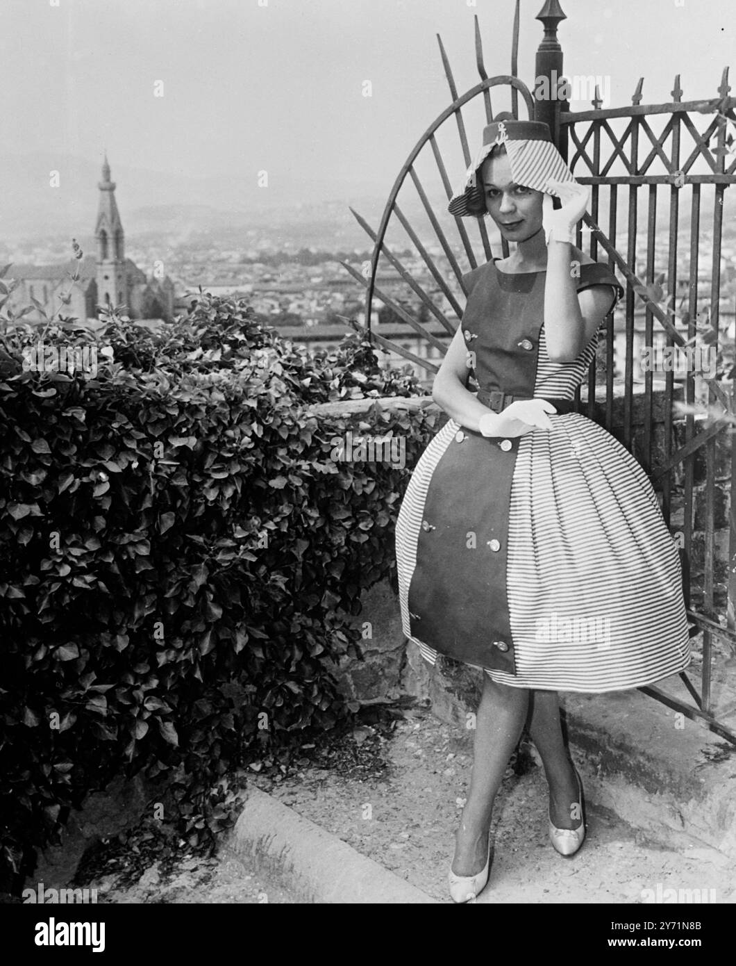 Modisches Falten. Florenz, Italien: Dieses Kleid von Vito of Mailand wurde dem Panorama von Florenz nachempfunden und ist aus blau gestreiftem weißem Baumwollelement mit Knopfverschluss gefertigt, das Schulterklappen bildet und Oberteil und Rock mit originalem Schürzeneffekt bedeckt. Der Rock ist ballonförmig und an den Seiten weich gefaltet, was eine Wellenbewegung zu den horizontalen Streifen ermöglicht. Das Kleid wird von einem Hut mit herabhängender Krempe und Ankerbesatz gekrönt, der aus den gleichen Materialien wie das Kleid hergestellt ist. Dieses Modell ist eines der Modelle, die während des Fashion Festivals in Florenz anlässlich der Präsentation der Herbst/Winter Kollektion 1959-60 durch die Leading gezeigt wurden Stockfoto