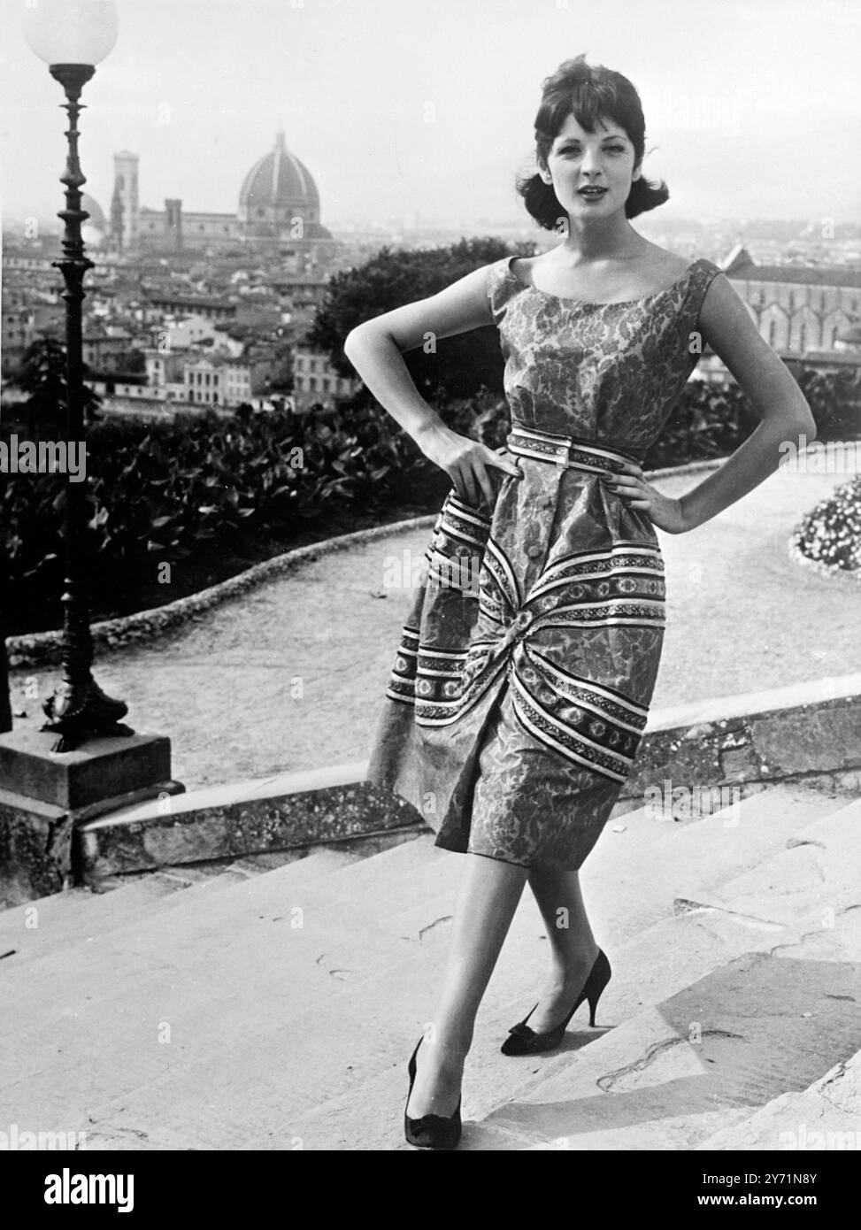 Modisches Falten. Florenz, Italien: Ein fröhliches Damastkleid mit roten Seidenapplikationen, das eine x-förmige Volute auf der Vorderseite des Rockes bildet, das dasselbe Design hat wie der dünne Gürtel, farbige Arabesken, die dem typischen Damastmuster des Kleides entsprechen, das ärmellos und mit einem ovalen Ausschnitt ist. Das Modell ist eines der Beroli-Kollektion für seine 1959-60er Mode im Florence Pitti Palace Show des italienischen Modus. 26. Juli 1959 Stockfoto