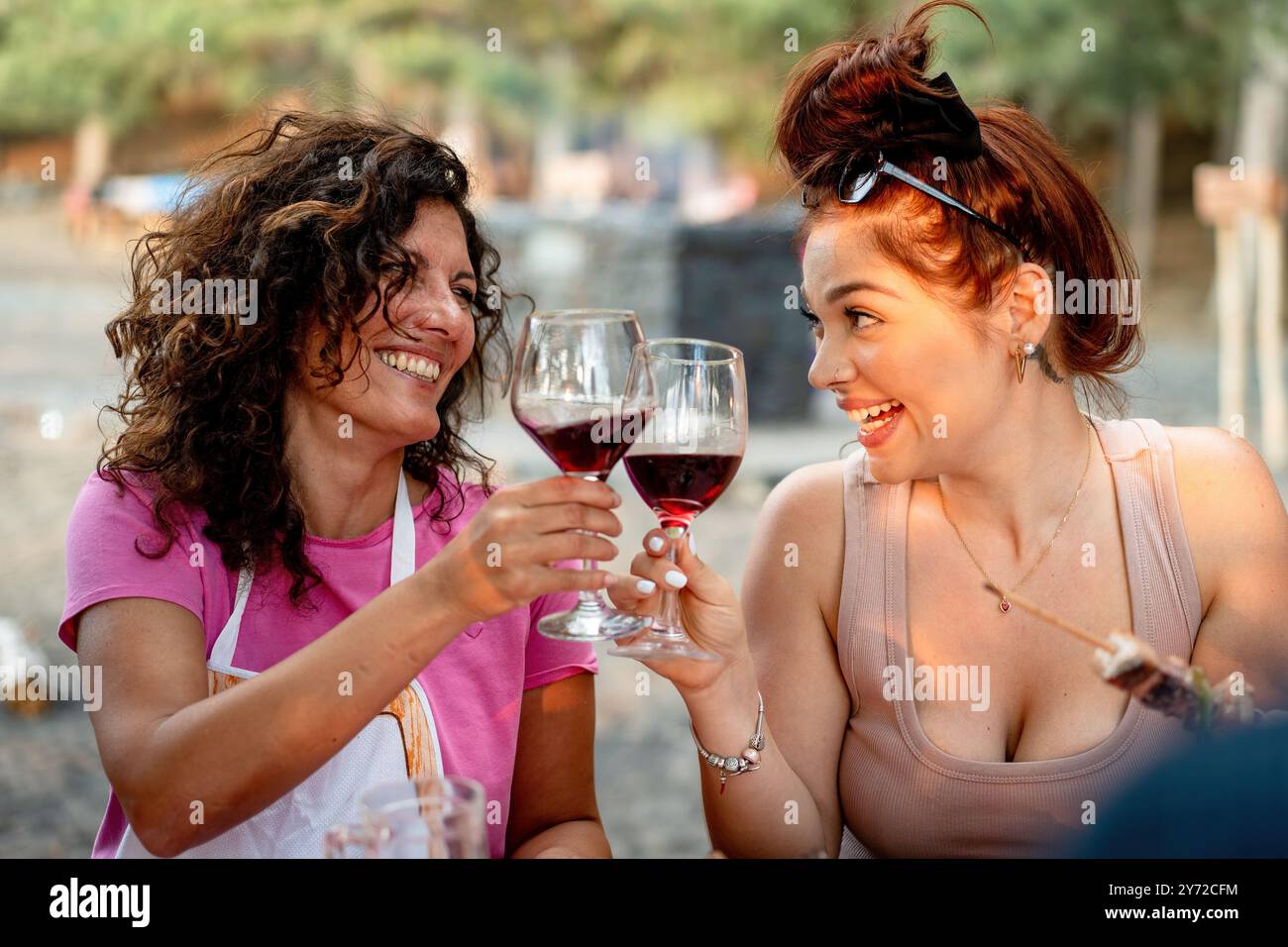 Zwei fröhliche Freundinnen, die bei einem Treffen im Freien mit Gläsern Rotwein tosten. Vielfältige Freundschaft, soziale Interaktion und Feier in einem nat Stockfoto