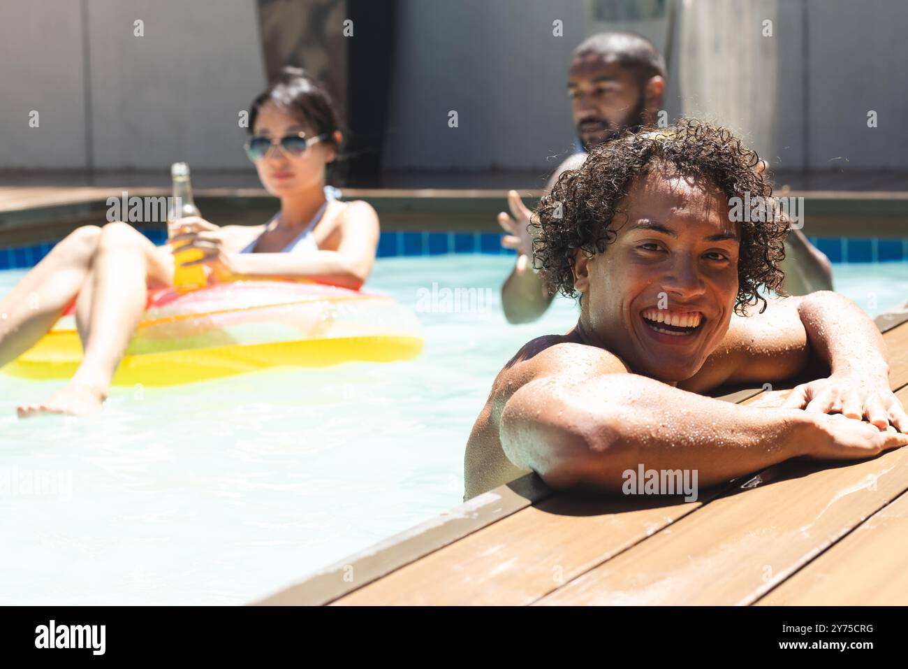 Lächelnder Mann im Pool mit verschiedenen Freunden, die sich auf dem Schwimmer entspannen und Bier trinken, zu Hause Stockfoto
