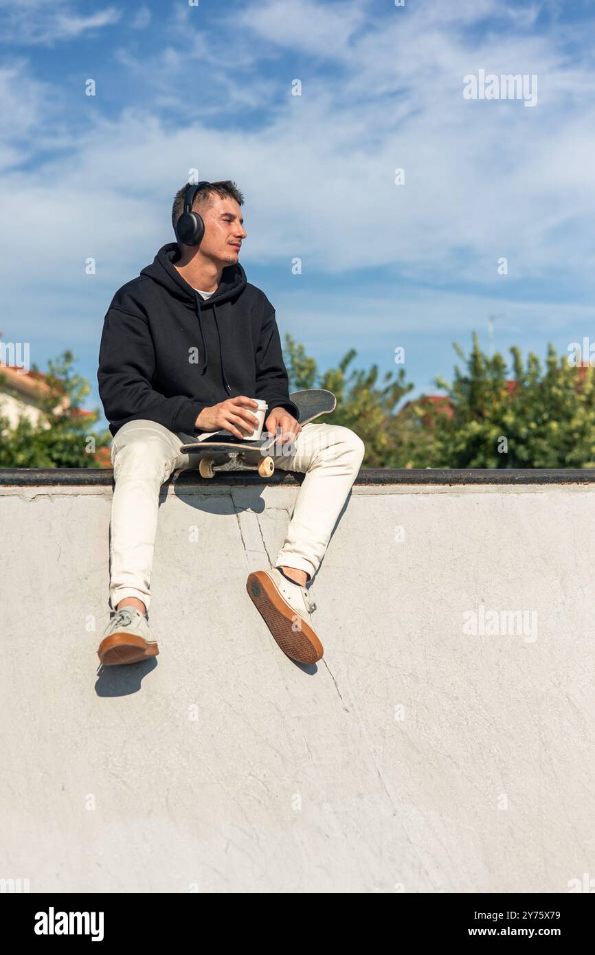 Vertikaler Skater sitzt am Rand einer Halfpipe, schlürft Kaffee und hört Musik über Kopfhörer. Verkörpert den Geist der Entspannung und en Stockfoto