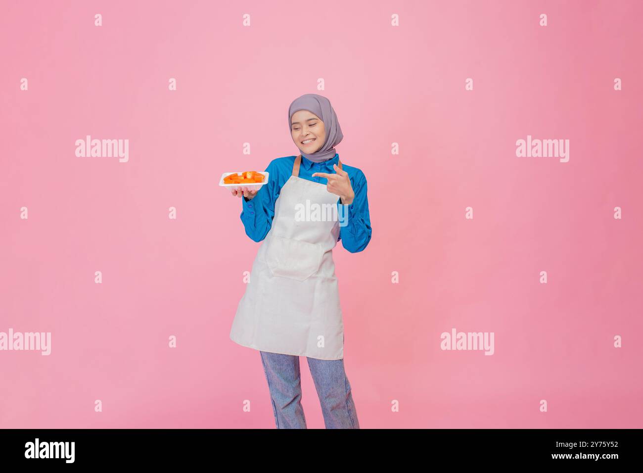 Eine fröhliche junge Frau in einer stilvollen Schürze hält eine Platte mit bunten Früchten vor einem rosafarbenen Hintergrund Stockfoto