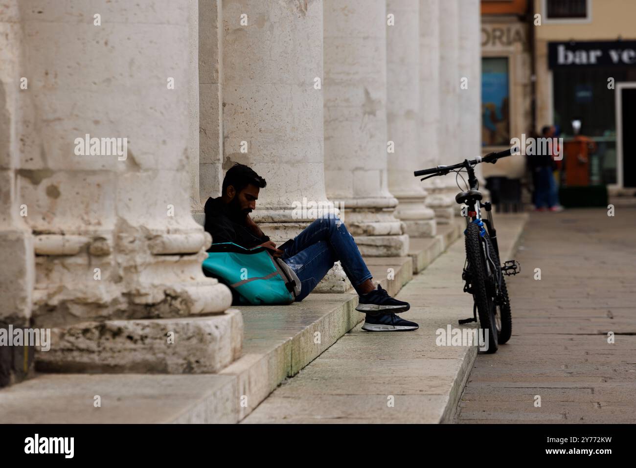 Vicenza, Italien - 13. September 2024: Ein Arbeiter aus Deliveroo macht eine Pause, sitzt auf der Treppe und lehnt sich an seine Arbeitstasche, während sein Fahrrad ruht Stockfoto