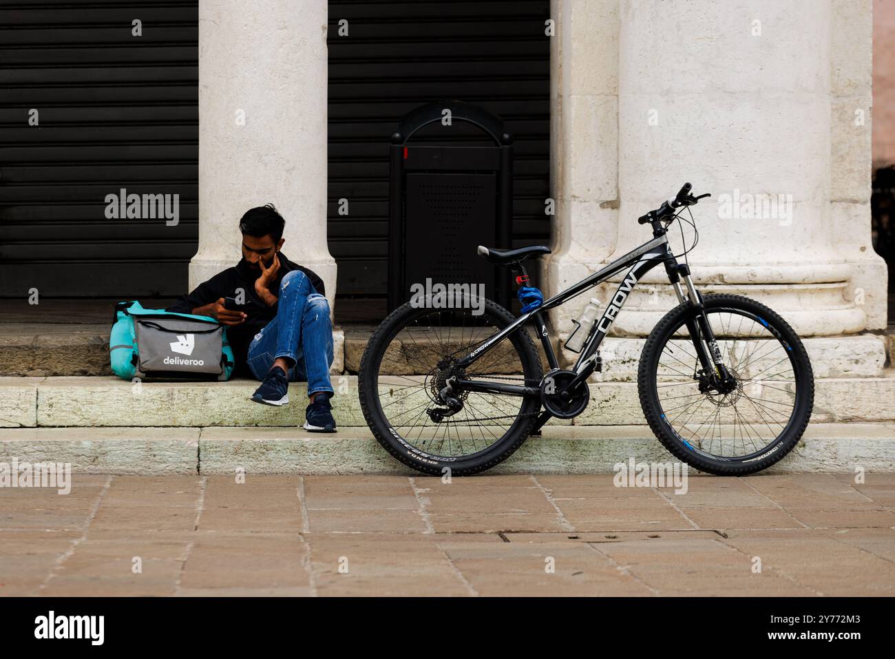 Vicenza, Italien - 13. September 2024: Ein Arbeiter aus Deliveroo macht eine Pause, sitzt auf der Treppe und lehnt sich an seine Arbeitstasche, während sein Fahrrad ruht Stockfoto